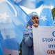 A woman in a blue hijab holds a sign reading Stop China's Uyghur Genocide behind blue flags representing East Turkestan.